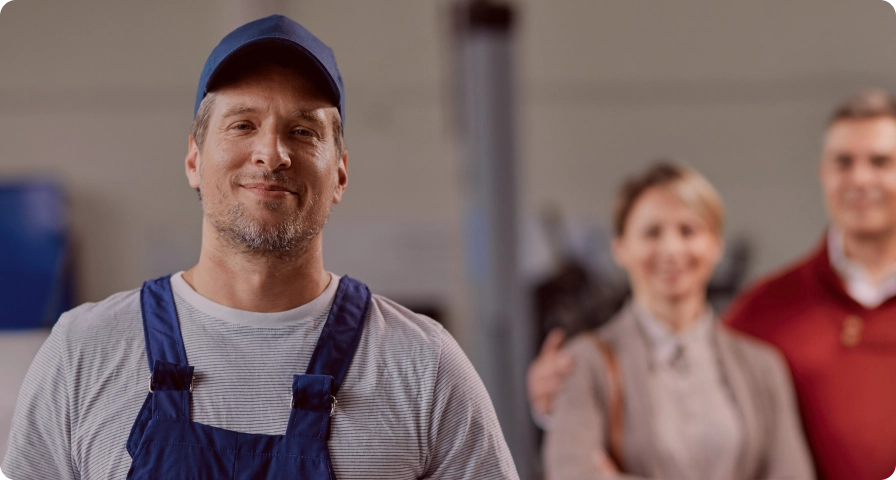Worker with blue basecap and working clothes looking into the camera. Mid age couple standing in the background smiling at the camera with blur effect.